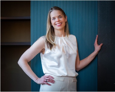 A picture of Caroline Butler stood in front of a teal background, smiling and with a hand on her hip and the other on the wall.