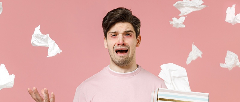 Sick unhealthy allergic man has red watery eyes, runny stuffy sore nose suffering from allergy trigger symptoms with paper napkin handkerchief isolated on pastel pink colour background.
