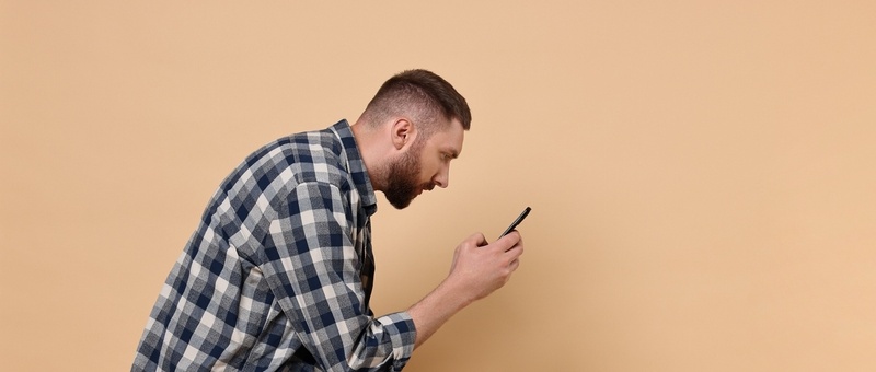 Man with poor posture sitting on chair and using smartphone against pale orange background.