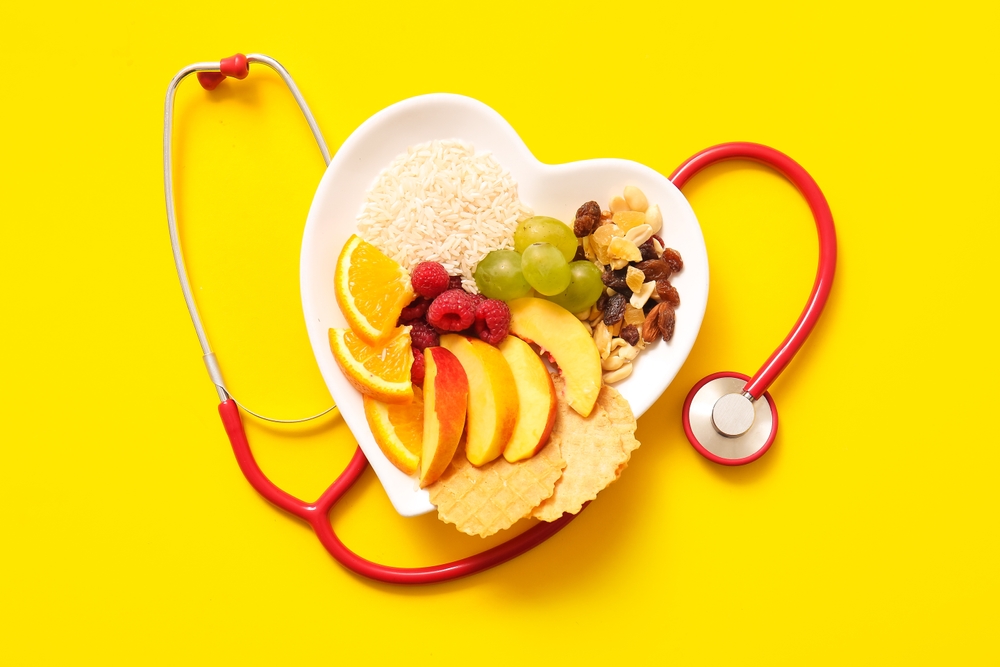 A heart-shaped white plate with fruit, rice, and nuts, surrounded by a red stethoscope on a bright yellow background.