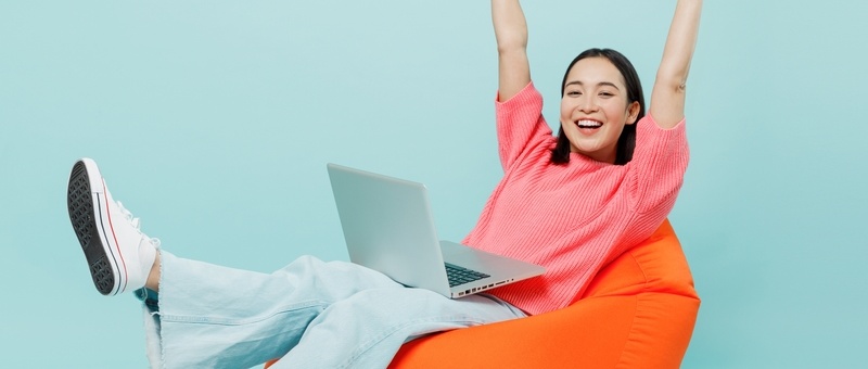 Young happy woman wearing pink jumper sitting in orange bag chair doing work on laptop computer with outstretched hands - isolated on pastel plain light blue background.