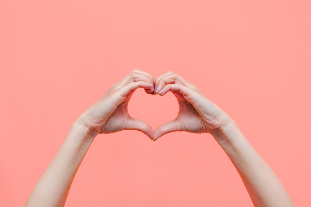 Pair of female hands in heart formation against a peach/orange/pink background.