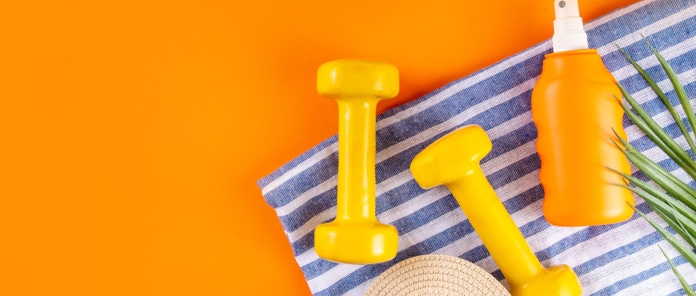 Summer sport and fitness activity background. Bright orange backdrop containing blue white striped beach towel, dumbbells for sports, sunscreen spray, summer hat and tropical palm leaves, copy space