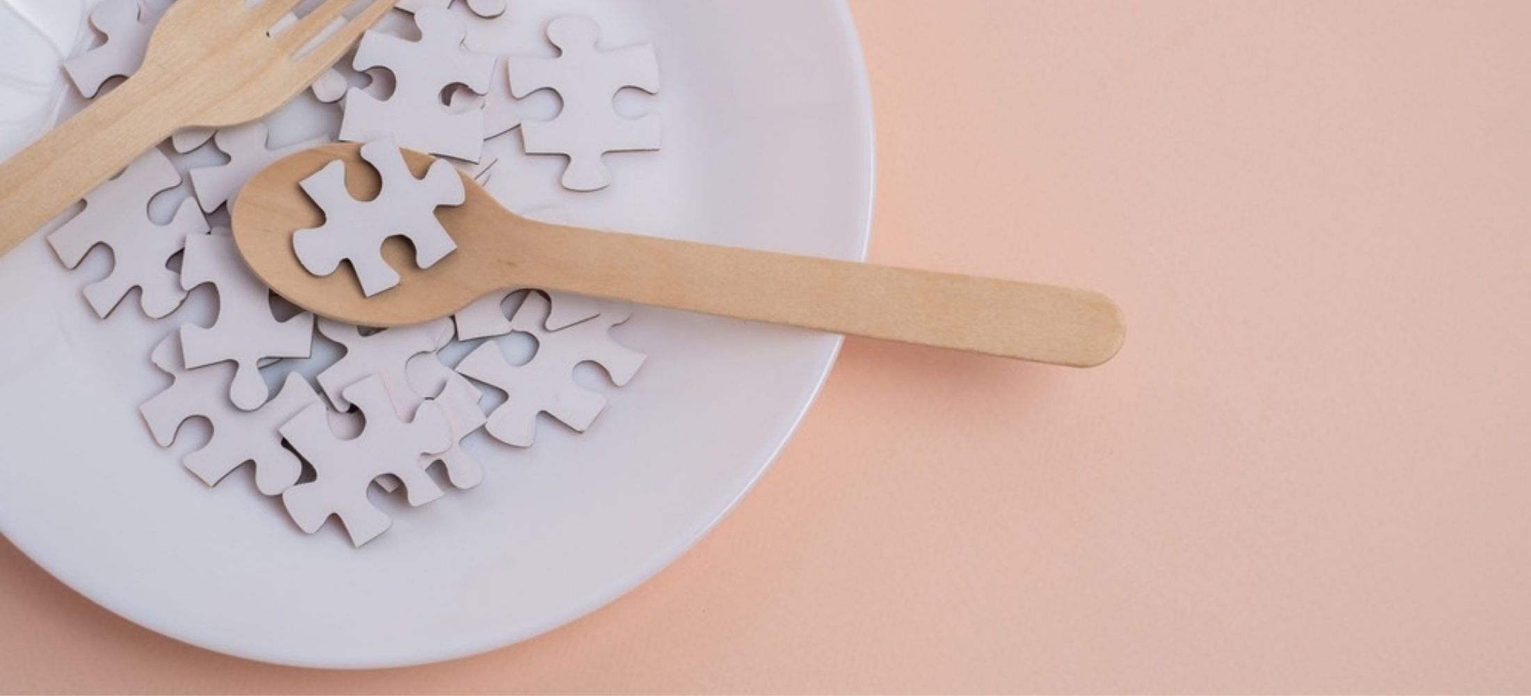 White jigsaw puzzle pieces on plate with wooden spoon and fork against pale peach background.