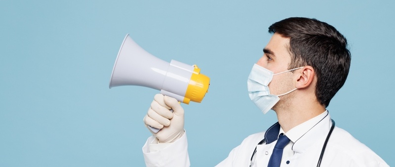 Young male doctor in white coat and face mask holding megaphone against blue background.
