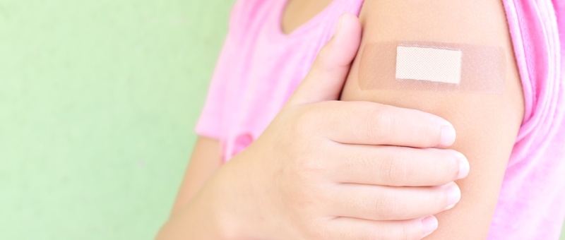Side view of woman in pink t-shirt holding arm with flu jab plaster against green background.