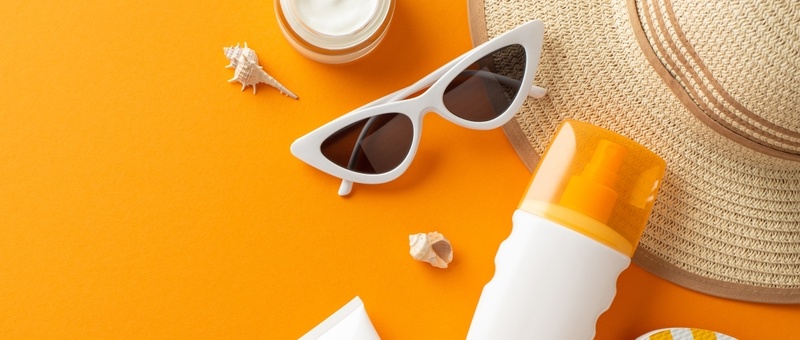 Top view photo of orange and white striped flip-flops, sunglasses, and straw hat, shells and sunscreen spray, tube and cream jar on orange isolated background.