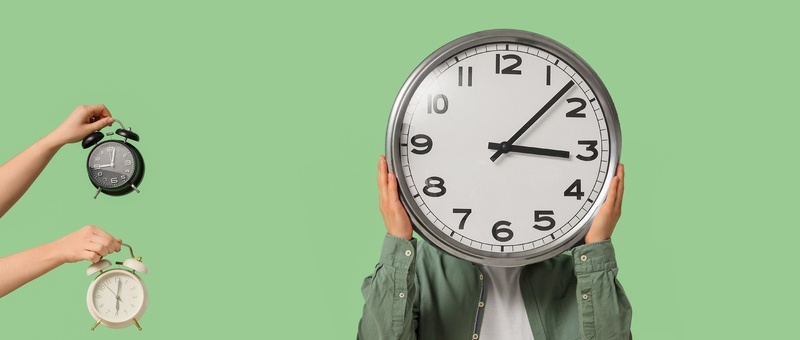 Person holding large clock in front of face with multiple alarm clocks held by hands on green background.