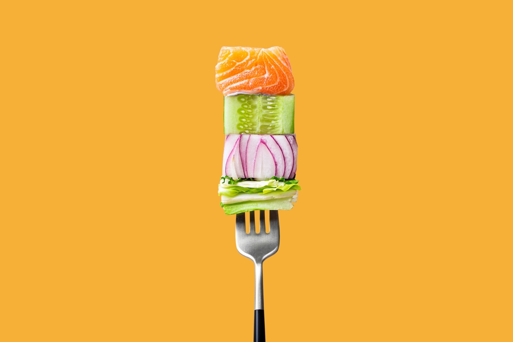 Stack of fresh ingredients on a fork: salmon, cucumber, red onion and lettuce against a bright orange background.