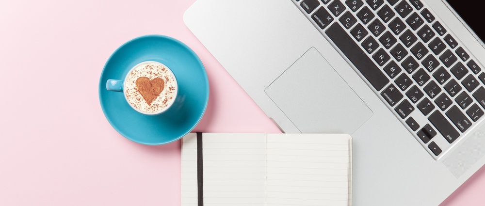 Cup of cappuccino with heart shape and laptop with note on pink background.