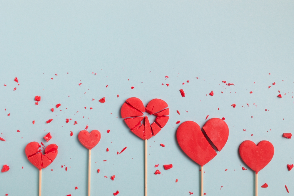 Five red heart-shaped lollipops in various stages of breaking apart, with scattered fragments, on a pale blue background, symbolising the physical symptoms of grief.