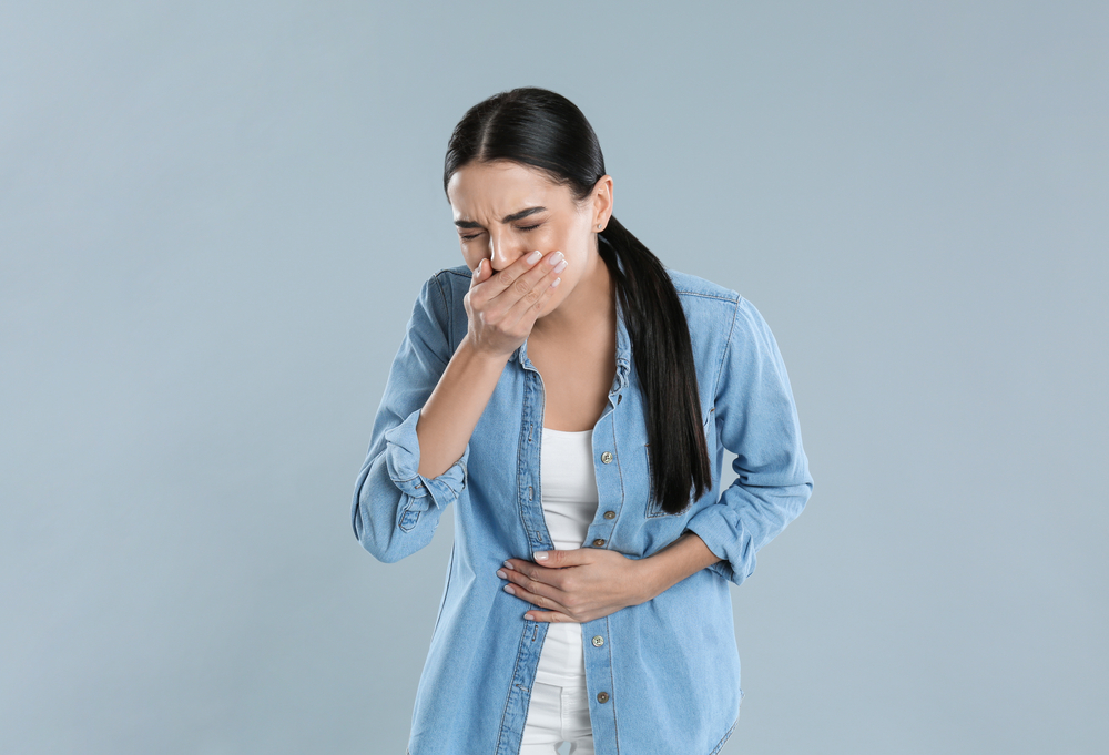 A young woman in a denim shirt covering her mouth and holding her stomach, appearing nauseous against a grey background.