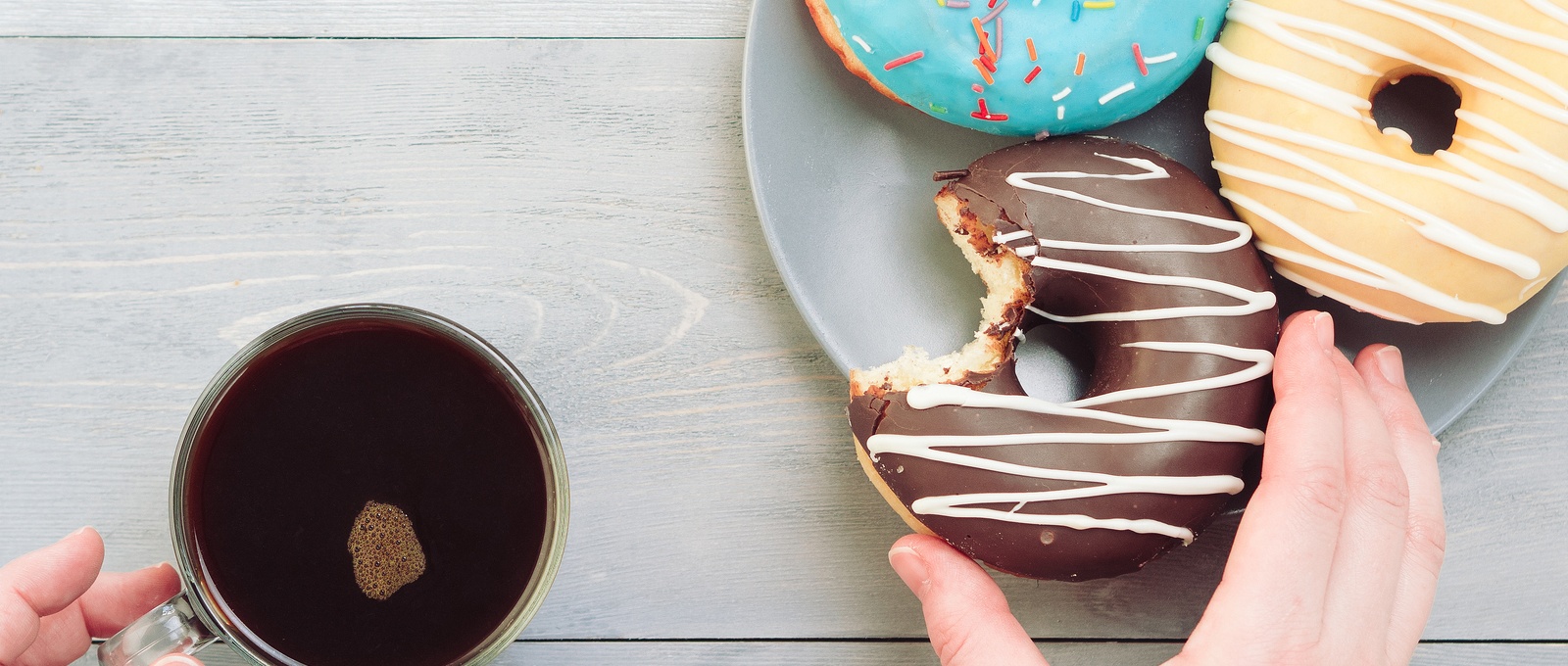 Woman hands with coffee and assorted doughnuts over grey wooden table. Top view of colourful doughnuts and coffee on grey wooden background.