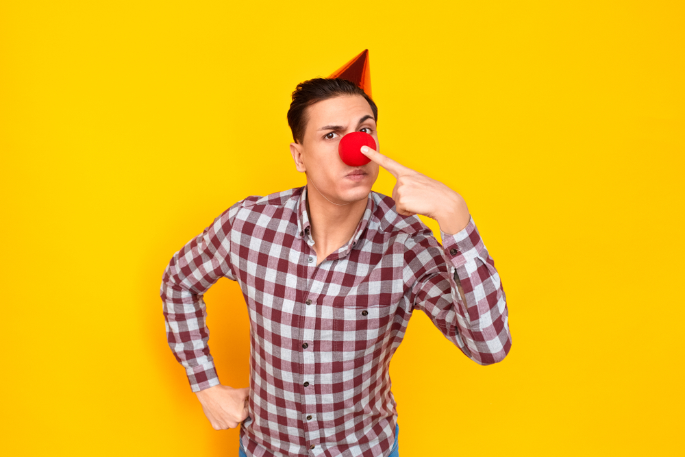 Clown man in party hat pointing finger at red nose against bright yellow background.