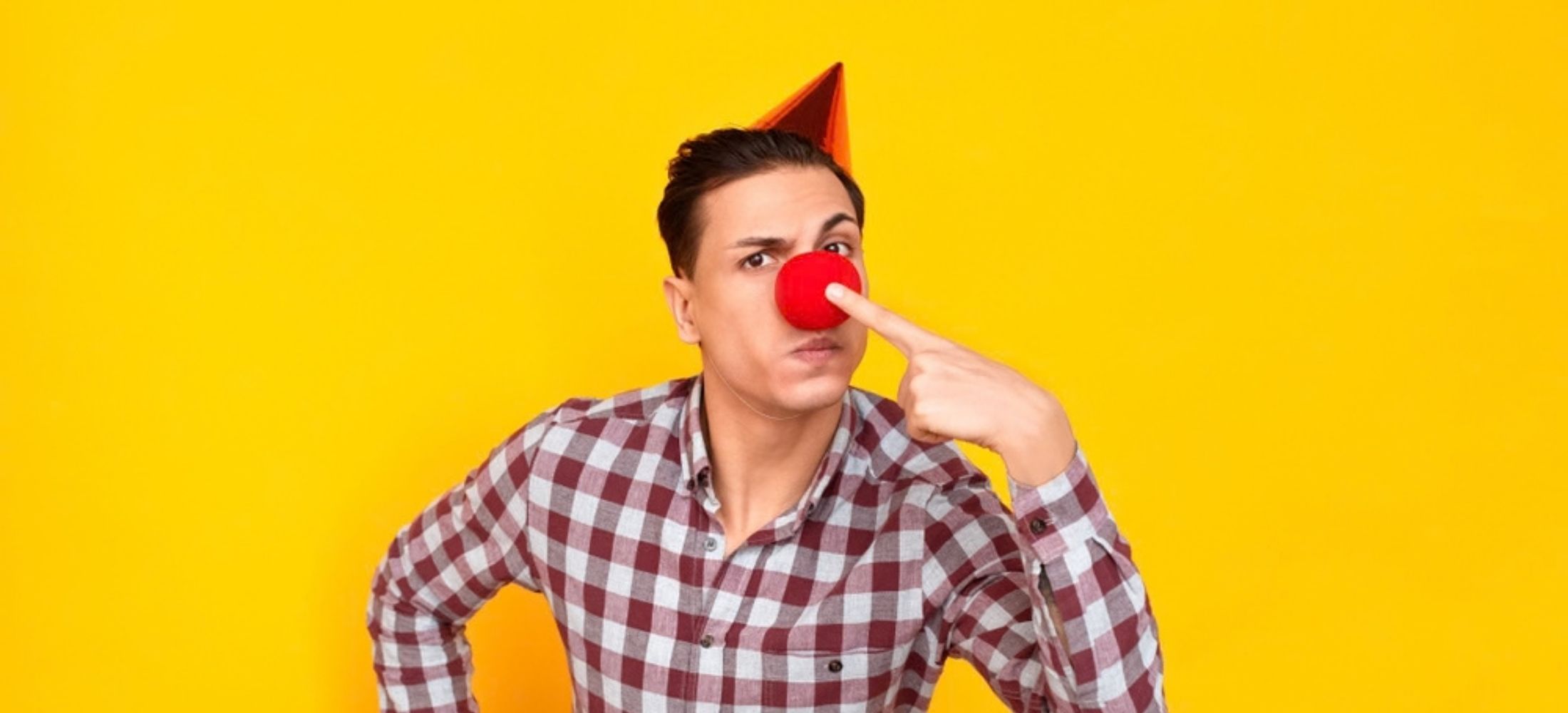 Clown man in party hat pointing finger at red nose against bright yellow background.