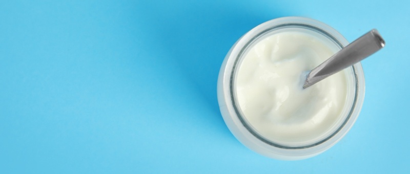 Organic Greek yoghurt in white bowl with silver spoon on light blue background, top view.