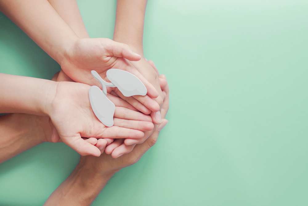 Two pairs of hands holding white paper lungs cut-out against pale gree background.