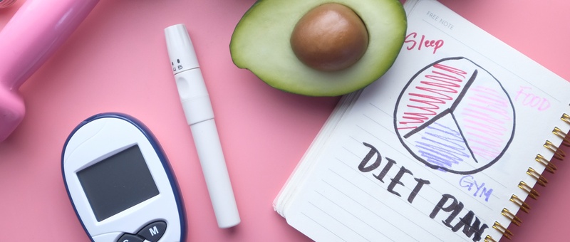 Avocado and notebook containing 'diet plan' diagram with blood sugar testing equipment and insulin pen against a pink background.