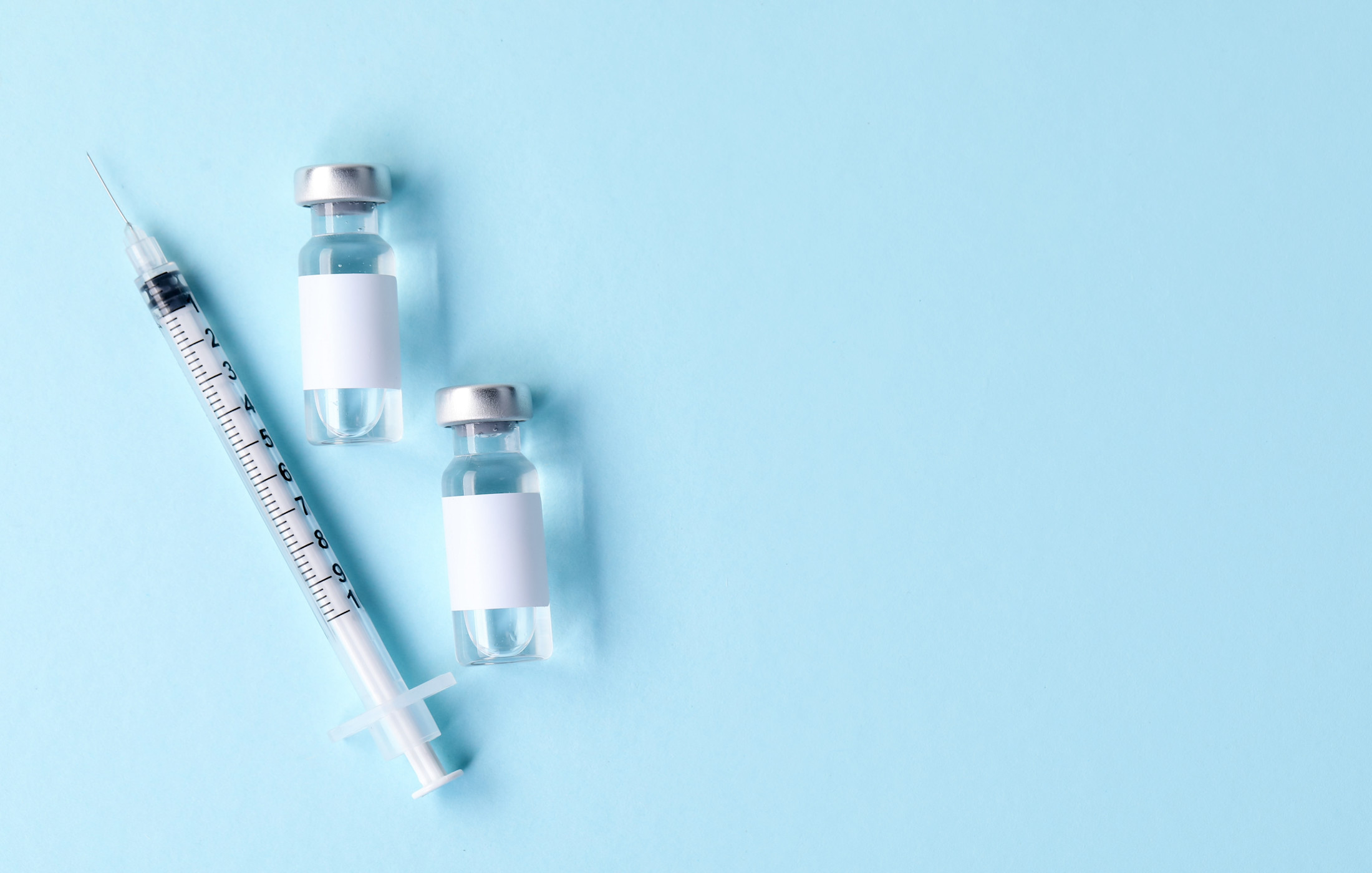 Medical syringe and two small glass vials with blank white labels arranged on a light blue background.