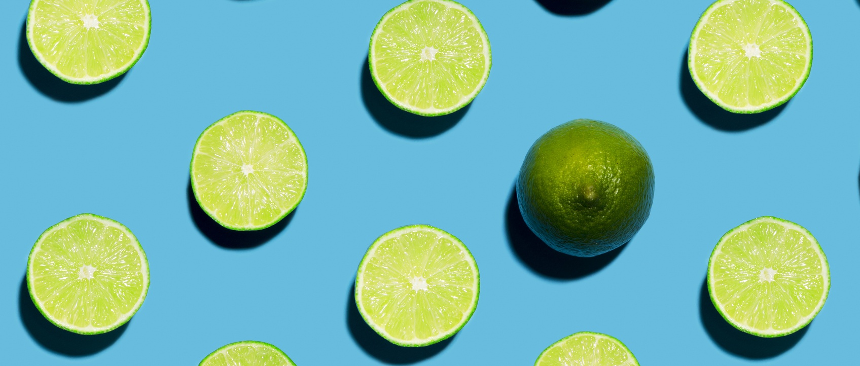 Fun brightly lit and colourful overhead photograph of sliced open green limes lined in rows with a harsh shadow on a blue background to understand juice cleanse.