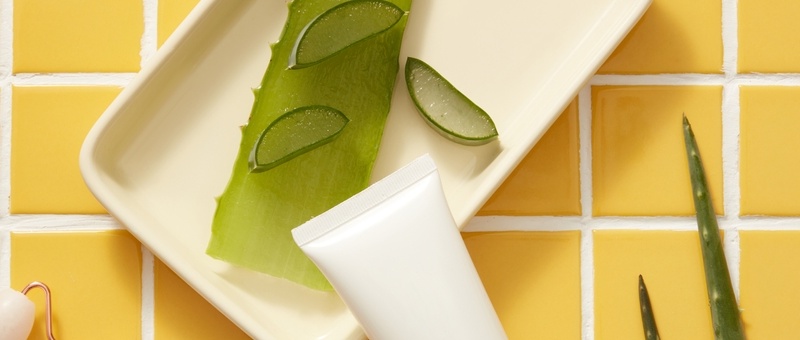 Green aloe vera stalks and white skin cream tube resting on white soap dish against yellow and white tiled bathroom backdrop.