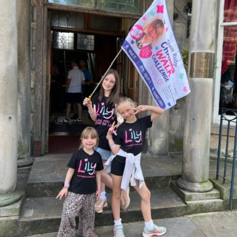 Ruby and her young sisters wearing Lily Foundation t-shirts and waving charity walk flag.