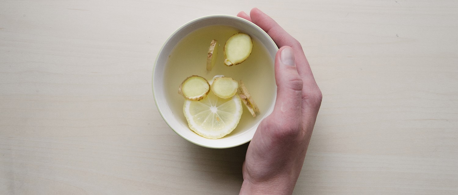 Hand holding white bowl of ginger and lemon tea with sliced ingredients floating in clear liquid.