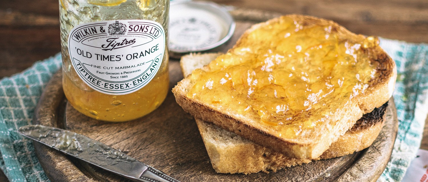 Toast spread with Wilkin & Sons 'Old Times' Orange marmalade on wooden board with jar and knife on teal cloth.