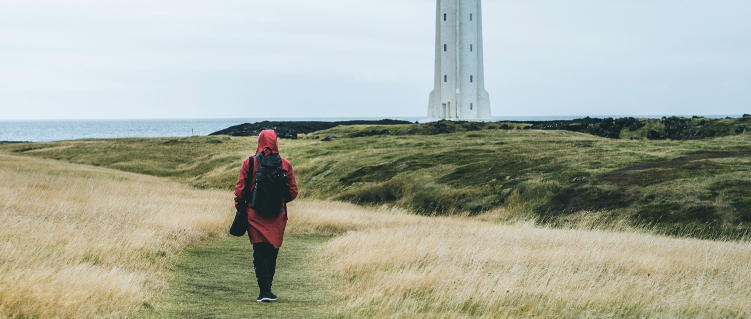 Person in red coat walking through coastal grassland towards a white lighthouse, with ocean in the background.