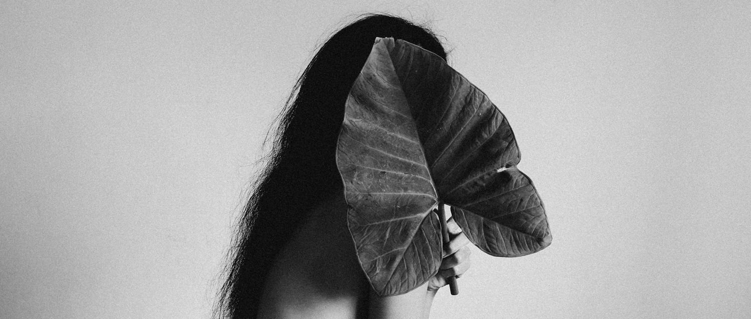 Black and white image of a person with long dark hair holding a large leaf that partially obscures their face.