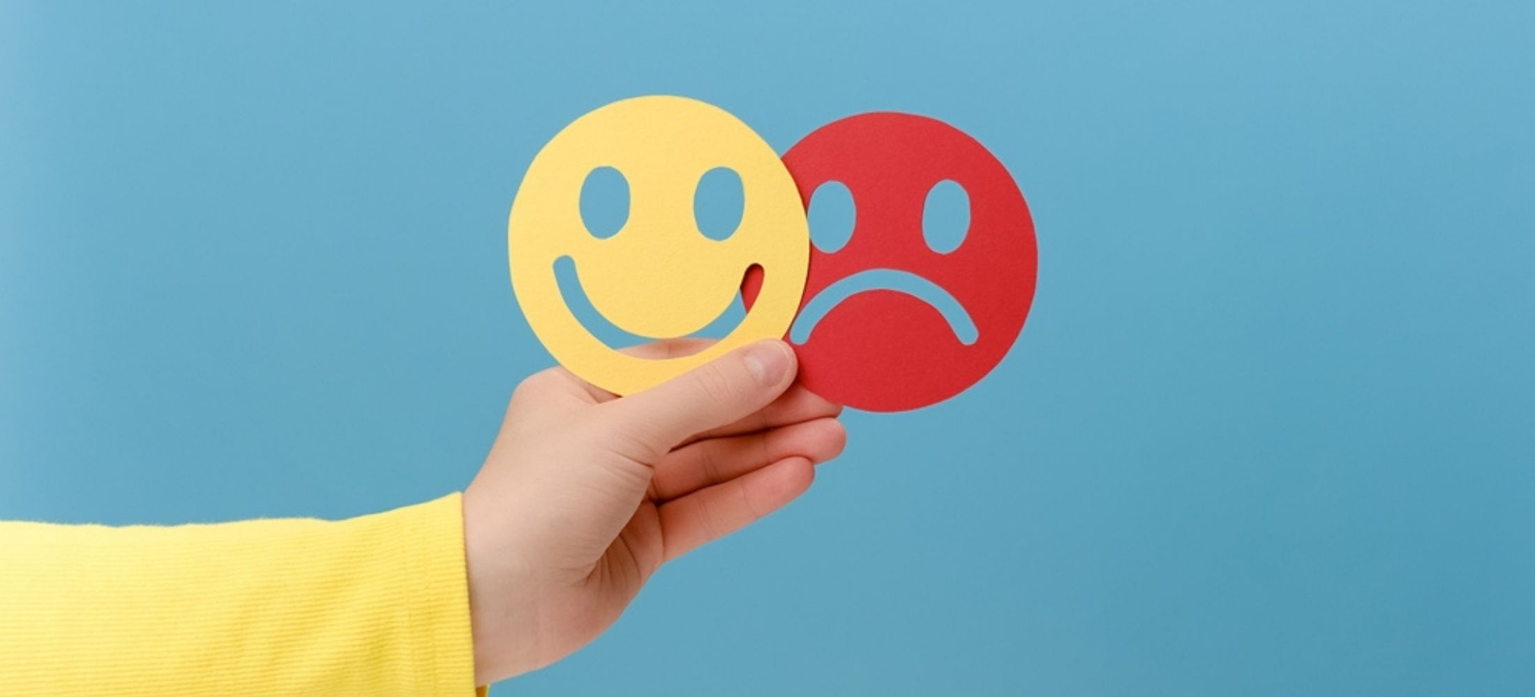 Close up of female hand holding happy and angry face paper, isolated on blue studio background.
