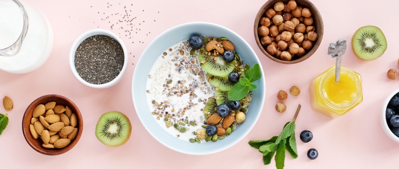 Homemade granola with milk, nuts, chia seeds, pumpkin seeds, honey, blueberries, and kiwi. Pink background.