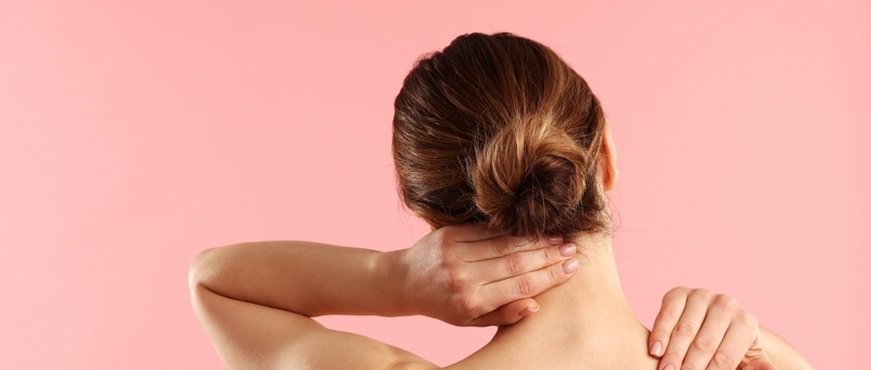 Woman with brown hair tied back, reaching behind to massage shoulders against a pink background.