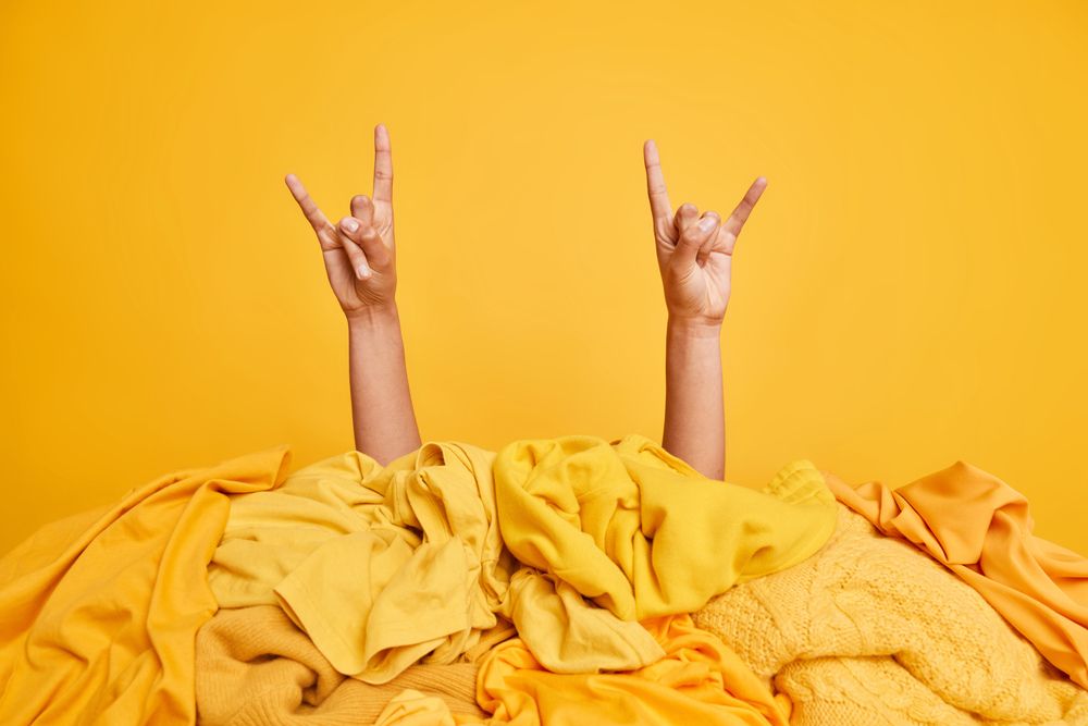Two arms rising out of yellow bed covers doing metal horns against yellow background.