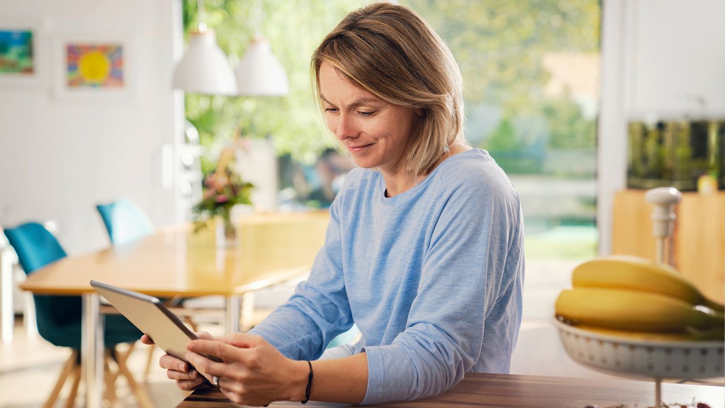 Eine Frau sitzt in einer hellen Küche am Tisch und bedient ein Tablet.