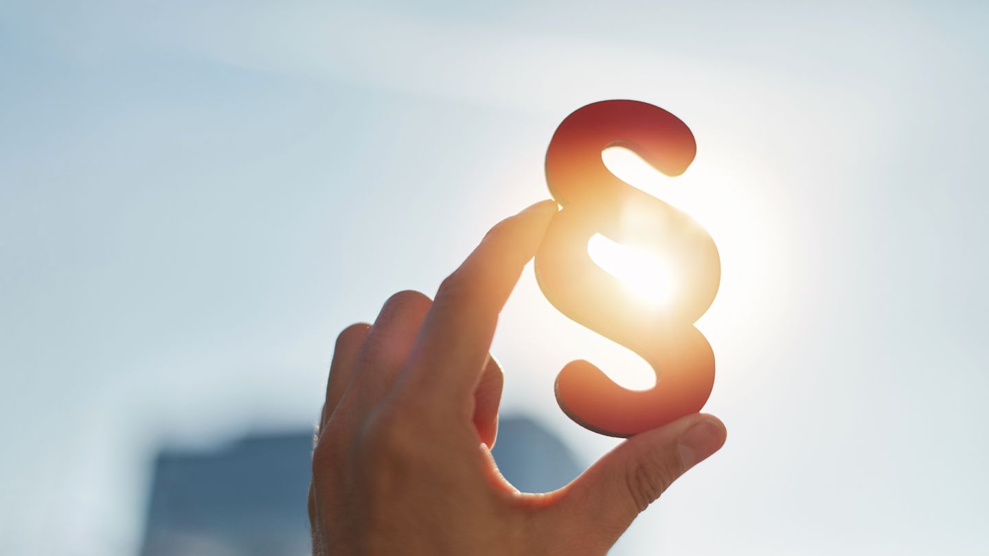 Eine Hand hält ein leuchtendes Paragraphen-Symbol gegen das Sonnenlicht.