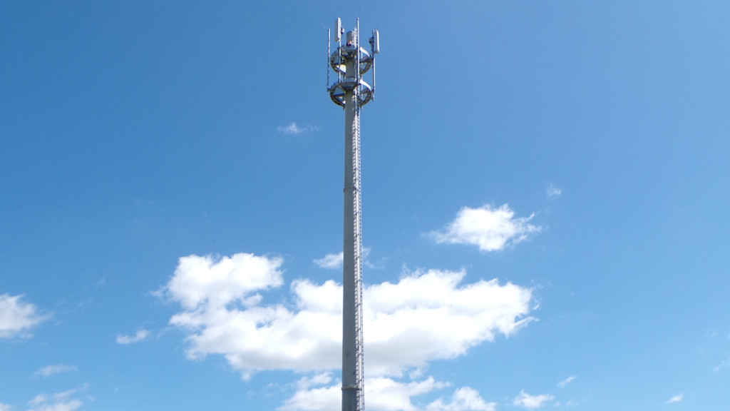 Hoher Funkmast auf einer Wiese unter blauem Himmel mit vereinzelten Wolken, im Vordergrund zwei geparkte Autos und eine Person in orangefarbener Arbeitskleidung am Mastfuß.