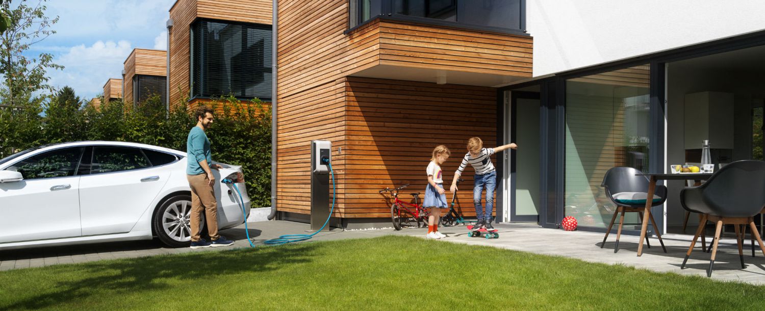 Ein Mann lädt ein Elektroauto an einer Wallbox vor einem modernen Einfamilienhaus auf, während zwei Kinder auf dem Gehweg spielen.