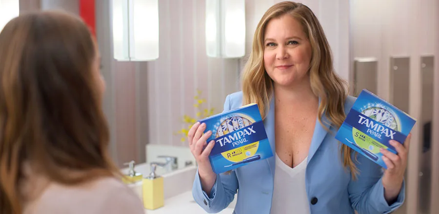 Amy Schumer standing in a blue jacket holding Tampax Pearl Regular and Tampax Pearl Super