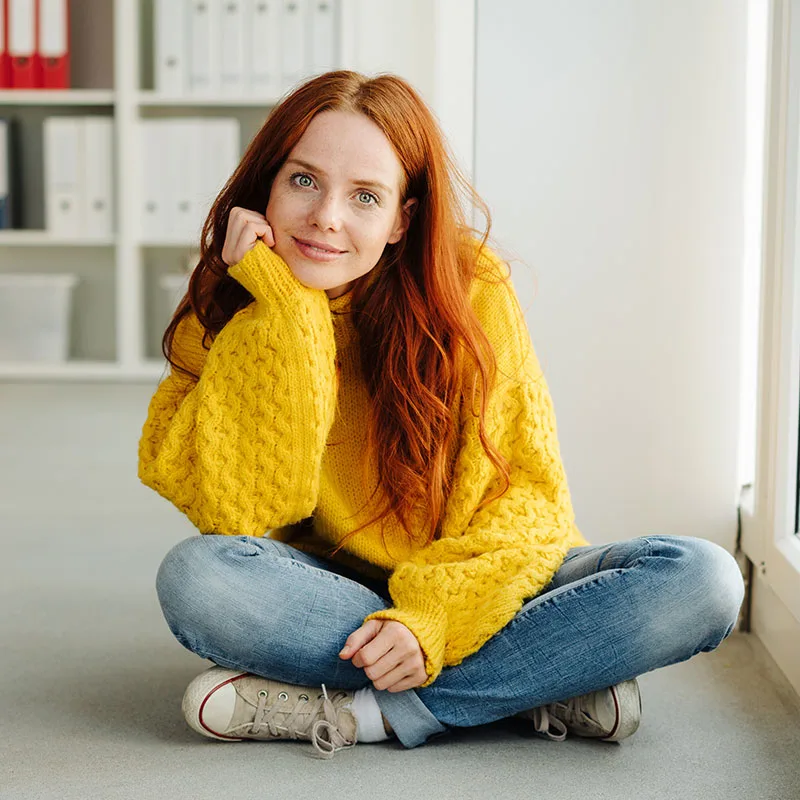 A young woman in a yellow sweater sitting on the floor in an office.