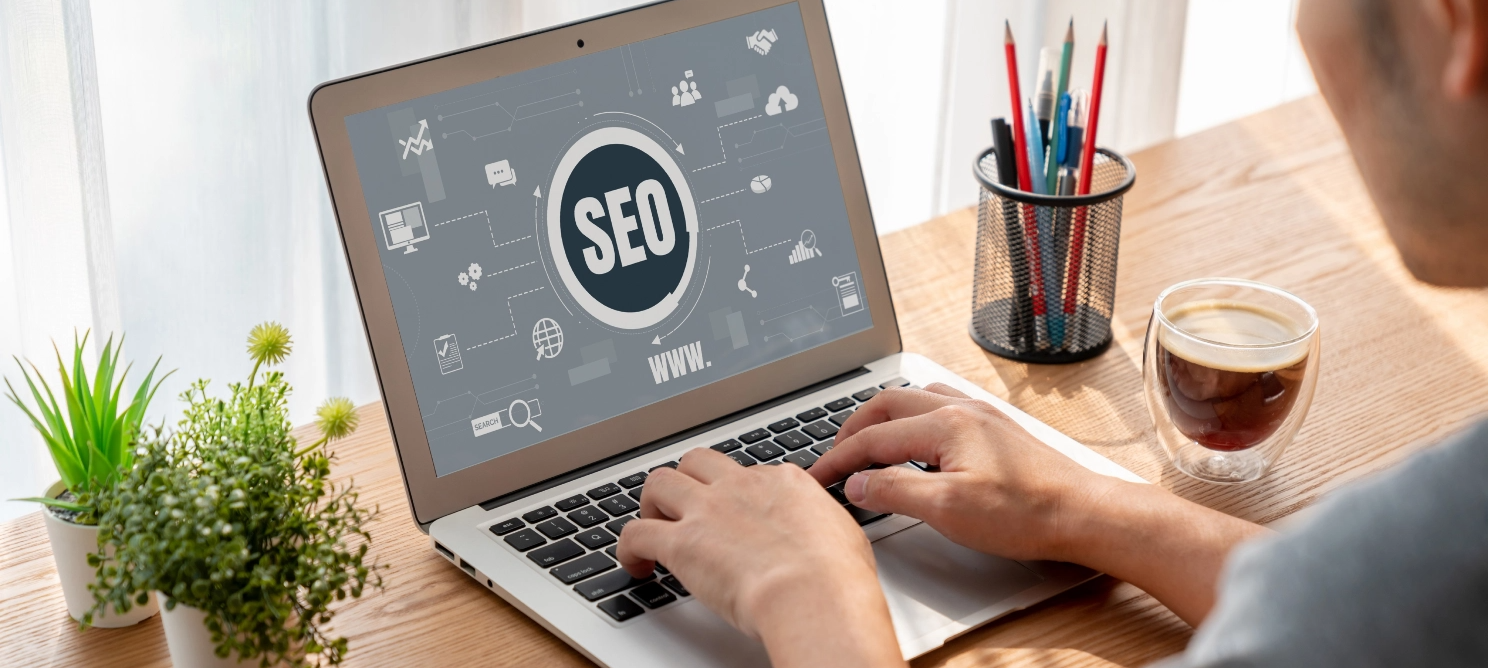 Person working on a laptop displaying a digital SEO interface on the screen, with a cup of coffee and pencil holder on the desk—illustrating search engine optimization in a business setting