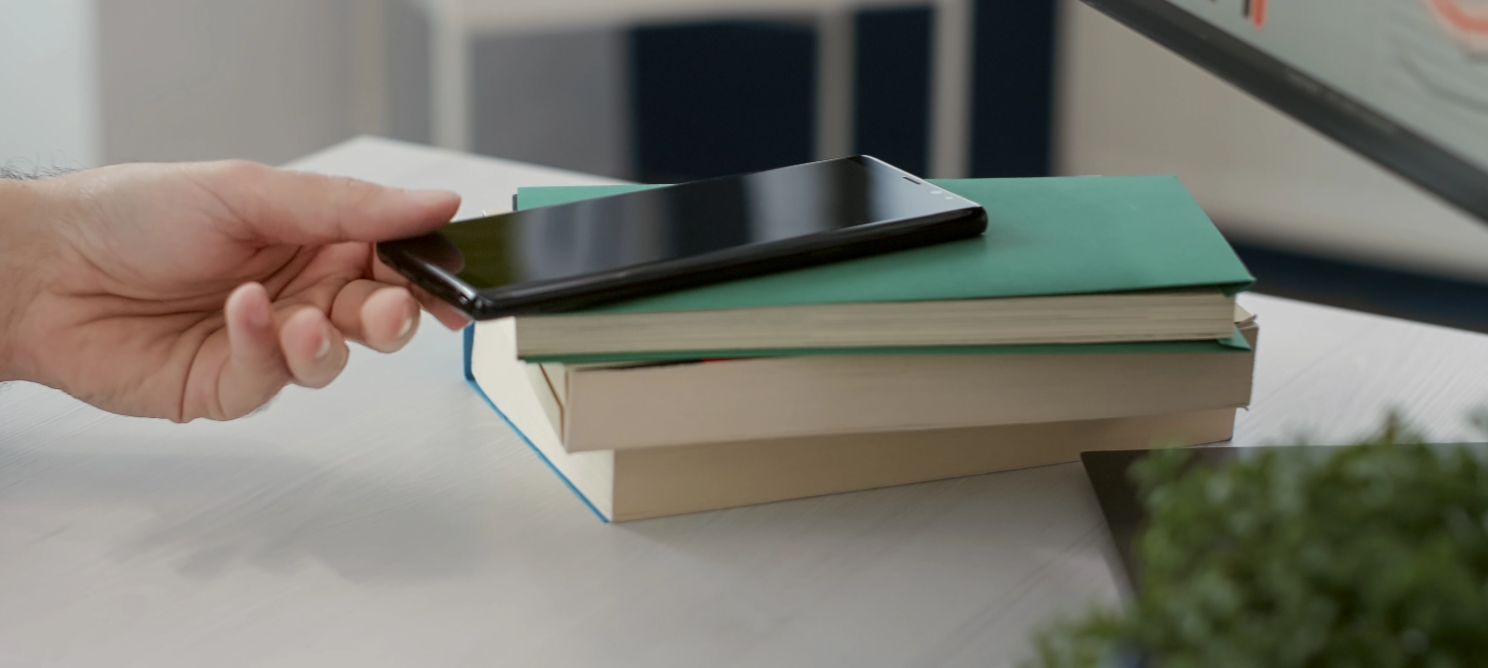 Person reaching for a smartphone placed on stacked books, symbolizing the use of mobile virtual interior design apps that enable users to visualize and customize home layouts