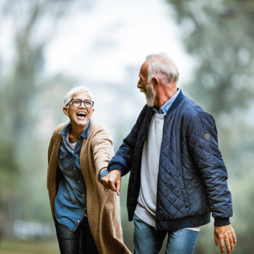 Pareja de adultos mayores sonrientes van cogidos de la mano. Ambos de pelo corto color blanco, la mujer viste una camisa de botones azul con un abrigo de color café y pantalón negro, el hombre viste un buso de color gris y chaqueta azul oscura.