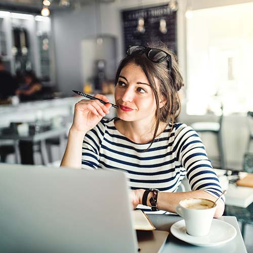 Mujer de pelo castaño recogido y lentes en la cabeza, luce buzo de rayas blancas y negras. Disfruta un café, al frente de su portátil y documentos