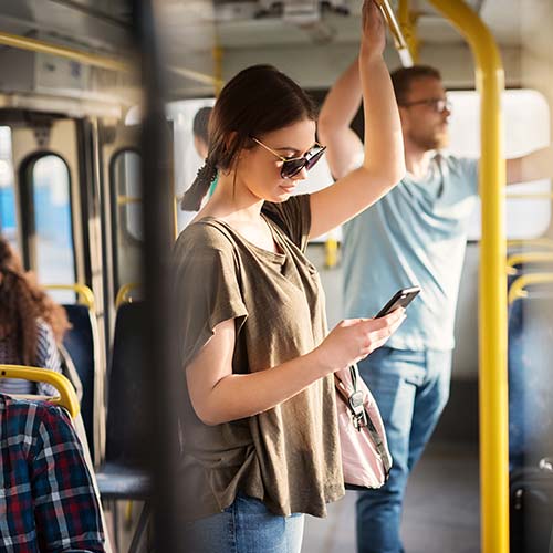 Parada en un autobús y sosteniéndose en un tubo amarillo, una mujer de pelo recogido, lentes y camiseta café, revisa su celular