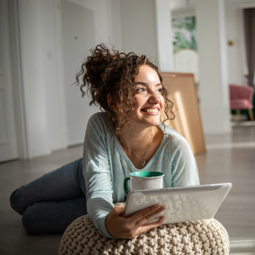 Mujer de pelo crespo y recogido, se encuentra en su apartamento sentada en el piso apoyando sus brazos en un puff color beige. Esta sonriente y pensativa sobre la planeación de su futuro para tener una mejor calidad de vida y bienestar financiero.