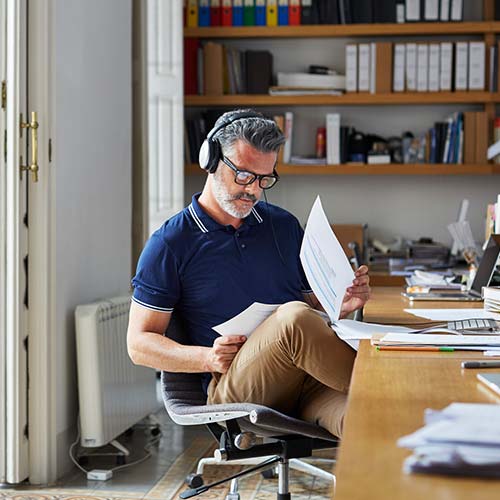 Hombre de pelo y barba cana, con lentes y audífonos, luce una camiseta polo azul oscura y pantalón café. Está revisando unos documentos