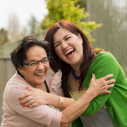 Madre e hija están abrazadas, sonrientes y felices. La madre es una señora adulta de pelo oscuro corto, gafas con marco negro y camisa rosa pastel. La hija de pelo oscuro esta vestida con camisa color gris y buso verde.