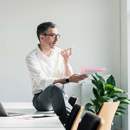 Sentado en un escritorio, hombre de pelo cano y lentes, luce camisa blanca. Habla por celular y sostiene unos documentos