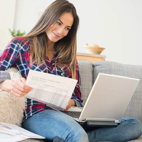 Sentada en un sofá gris, mujer de pelo castaño con camisa de cuadros, lee en su portátil y sostiene un documento